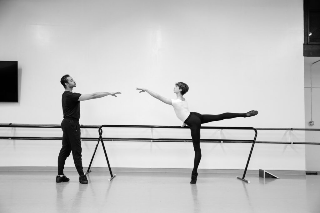 In this black and white photo, Adrian Blake Mitchell coaches a teenage male ballet student as he practices second arabesque on relevé at the barre. The boy, in black tights and ballet slippers and a white t-shirt, balances on his left leg. Black Mitchell, wearing a black t-shirt, pants and black sneakers, stands in front of him at the barre, facing the student, and reaches his right arm out to match his line.