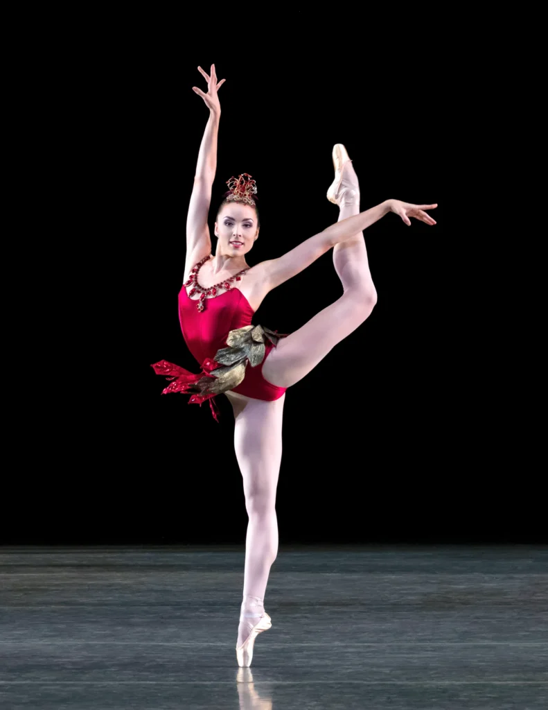Emily Kikta, wearing a red bejeweled costume with a short, paneled skirt and tall tiara with red rhinestones, does a high grand battement on pointe with her her back leg in a turned-in attitude. Her back foot is higher than her head. She raises her arms and looks out confidently towards the audience with a sultry smile.