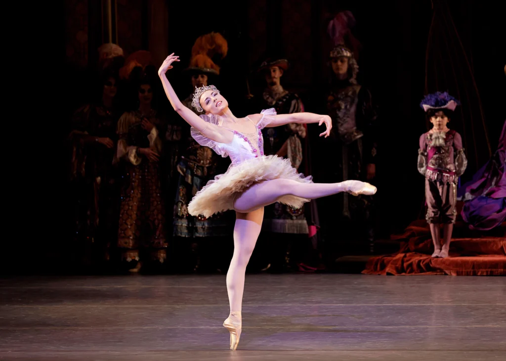 Emily Kikta does a piqué attitude devant onstage during a performance, raising her right leg. She arches back slightly with her arms in third position and projects out dreamily to the audience. She wears a light purple tutu and large rhinestone tiara, pink tights and pointe shoes. A group of dancers costumed as medieval courtiers and pages stand in the background and watch.