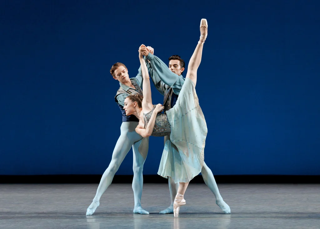 Emily Kikta dances with two male dancers onstage, all of them costumed in light blue. She does a deep penché on pointe with her left leg raised as one man holds her left arm up. She wraps her right arm around her ribcage, holding the hand of the other dancer as he threads his arm through. Both men stand behind her and facing each other in tendu, wearing blue tights and ballet slippers, blue vests with blue-green collared shirts underneath. Kikta wears a knee-length blue dress with think straps and a blue-and-gold brocade bodice.