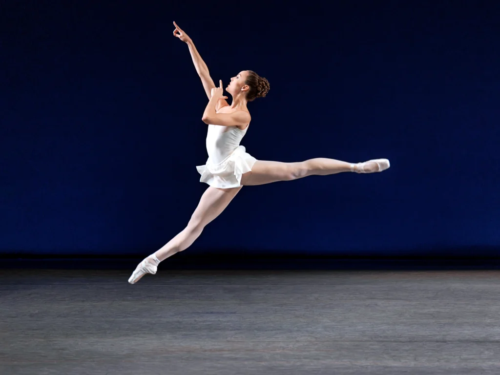 Emily Kikta, wearing a white leotard, white skirt, pink tights and pointe shoes, does a giant sissone en avant onstage during a performance. She holds her right arm up in front of her, pointing her index finger, and pulls her other pointed index finger close to her mouth.