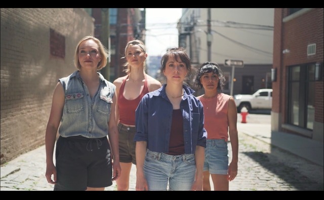A group of four female dancers wearing shorts and short-sleeved shirts, stand in the middle of a city alley on a sunny day and look out beyond the camera.