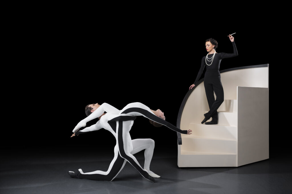 In front of a pitch black background, two dancers mirror each other's positions in a forward lunge, crossing one over the other's lines. One wears a white unitard, and the other white with a black stripe down the sides. Behind them to the right, a dancer in a black suit stands on a curved white staircase.