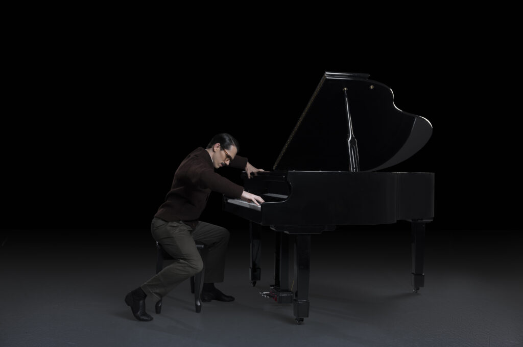 A man in dark clothing, portraying Igor Stravinsky, hunches over a black grand piano.