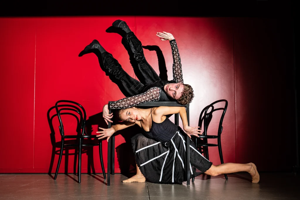 A male and female dancer pose with a black café table in front of a bright red wall. The woman lunges deeply under the table, stretching her left leg out behind her as she presses her head against the table. She wears a black dress with white stripes on the skirt. The man lays on his back on top of the table and lifts his legs up, his feet flexed, while raising his right arm up and turning his wrist in. He wears black pants, and white and black patterned shirt and black ballet slippers. Both dancers look towards the camera.