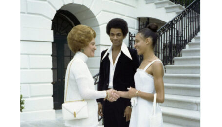 First Lady Betty Ford shakes hands and smiles with Sandra Fortune-Green at the base of the front steps leading up to the White House. Ford wears a white turtleneck, while Fortune-Green wears a white dress, and both carry white purses. Sylvester Campbell, wearing a black suit with an open collared-shirt, stands next to Fortune-Green on her right side and smiles at Ford.
