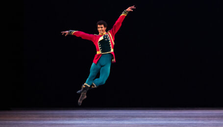 Gian Carlo Perez jumps up into a jeté onstage during a performance of Stars and Stripes, with his left leg in coupé derrierre. He wears a red jacket with gold and dark blue trim, bright blue tights and black boots. His arms are open to the side, his left arm slightly higher than the right, and he looks directly towards the audience and smiles.