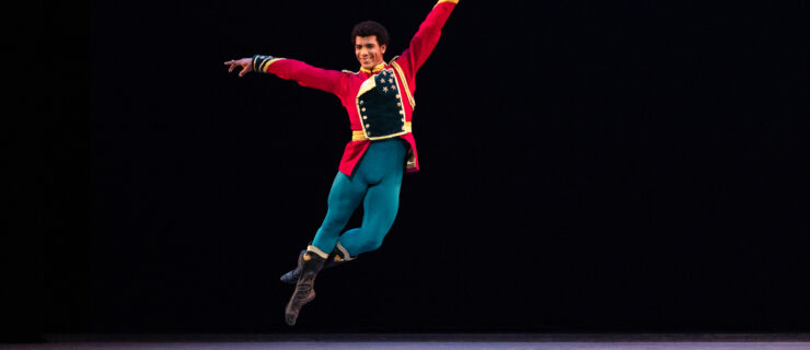 Gian Carlo Perez jumps up into a jeté onstage during a performance of Stars and Stripes, with his left leg in coupé derrierre. He wears a red jacket with gold and dark blue trim, bright blue tights and black boots. His arms are open to the side, his left arm slightly higher than the right, and he looks directly towards the audience and smiles.