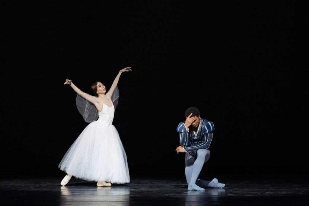 Yuriko Kajiya and Gian Carlo Perez portray the characters of Gielle and ALbrecht, respectively, in a performance of the ballet Giselle. Kajiya wears a white romantic tutu and stands in tendu derriere, lifting her amrs up in a high V-shape. Perez, in a blue tunic, tights and ballet slippers, kneels down onto his right knee and holds his head in his right hand.