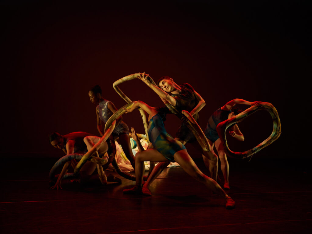 Onstage, lit only by yellow and red from a central light source, a group of dancers in shiny unitards cluster together around a decorative snake-like art installation.