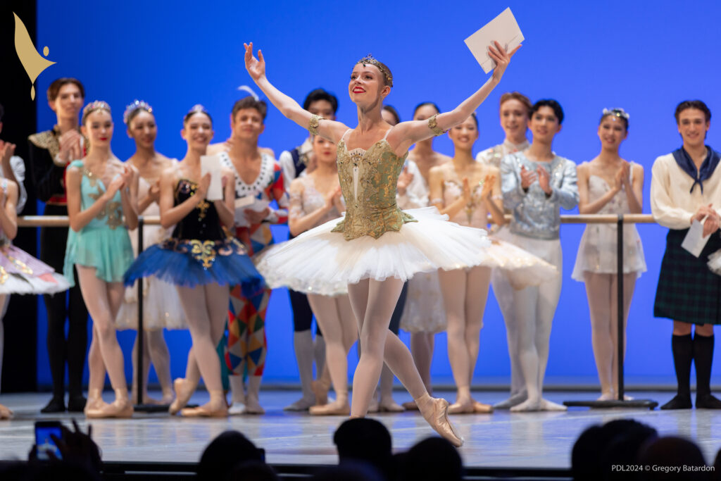 Ruby Day, wearing a gold and white tutu, pink tights, pointe shoes and a gold headpiece, stands at the front of a stage in B plus and opens her arms wide to the audience. She has a happy smile on her face. A group of other teenage male and female dancers in various classical ballet costumes stand behind him upstage and clap.