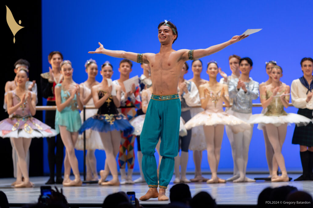 Martinho Lima Santos, wearing blue harem pants, ballet slippers and a blue headband with feather, stands at the front of a stage and opens his arms wide to the audience. He has a huge smile on his face. A group of other teenage male and female dancers in various classical ballet costumes stand behind him upstage and clap.