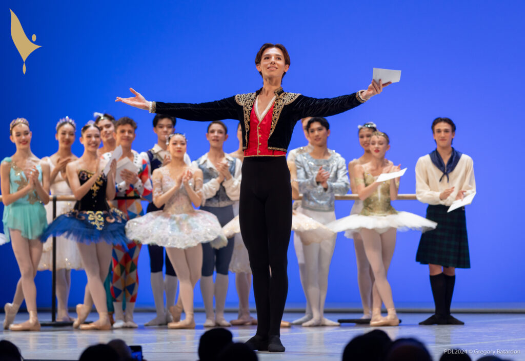 Juliann Fedele-Malard, wearing black tights and ballet slippers, a red vest and a black bolero jacket with gold trim, stands at the front of a stage and opens his arms wide to the audience. He has a smile on his face. A group of other teenage male and female dancers in various classical ballet costumes stand behind him upstage and clap.