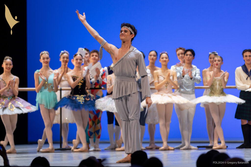 Jenson Blight, wearing a gray harem pants, a one-sleeved gray shirt gathered at the waist, and tan ballet slippers, stands at the front of a stage and lifts his right arm in preparation for a bow. He out into the audience and smiles. A group of other teenage male and female dancers in various classical ballet costumes stand behind him upstage and clap.