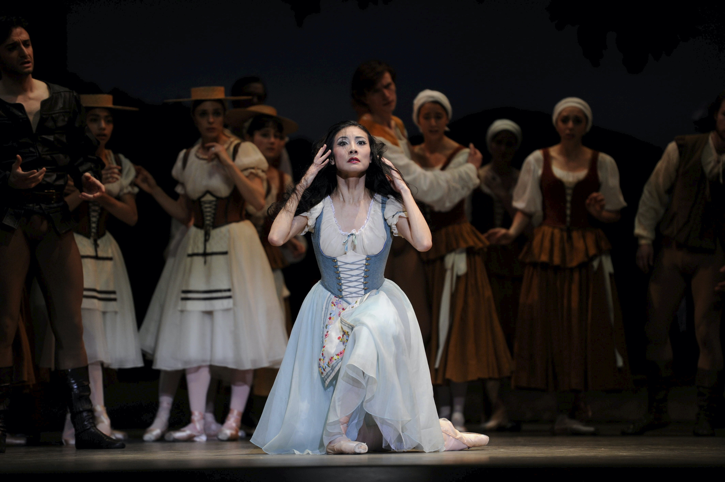 During a performance of Giselle, Yuan Yuan Tan, wearing a blue and white peasant dress, kneels down and touches her face. She looks out into the distance, her long black hairdown and messy. A large corps of male and female dancers in peasant costumes stand behind her, watching with concerned looks on their faces.