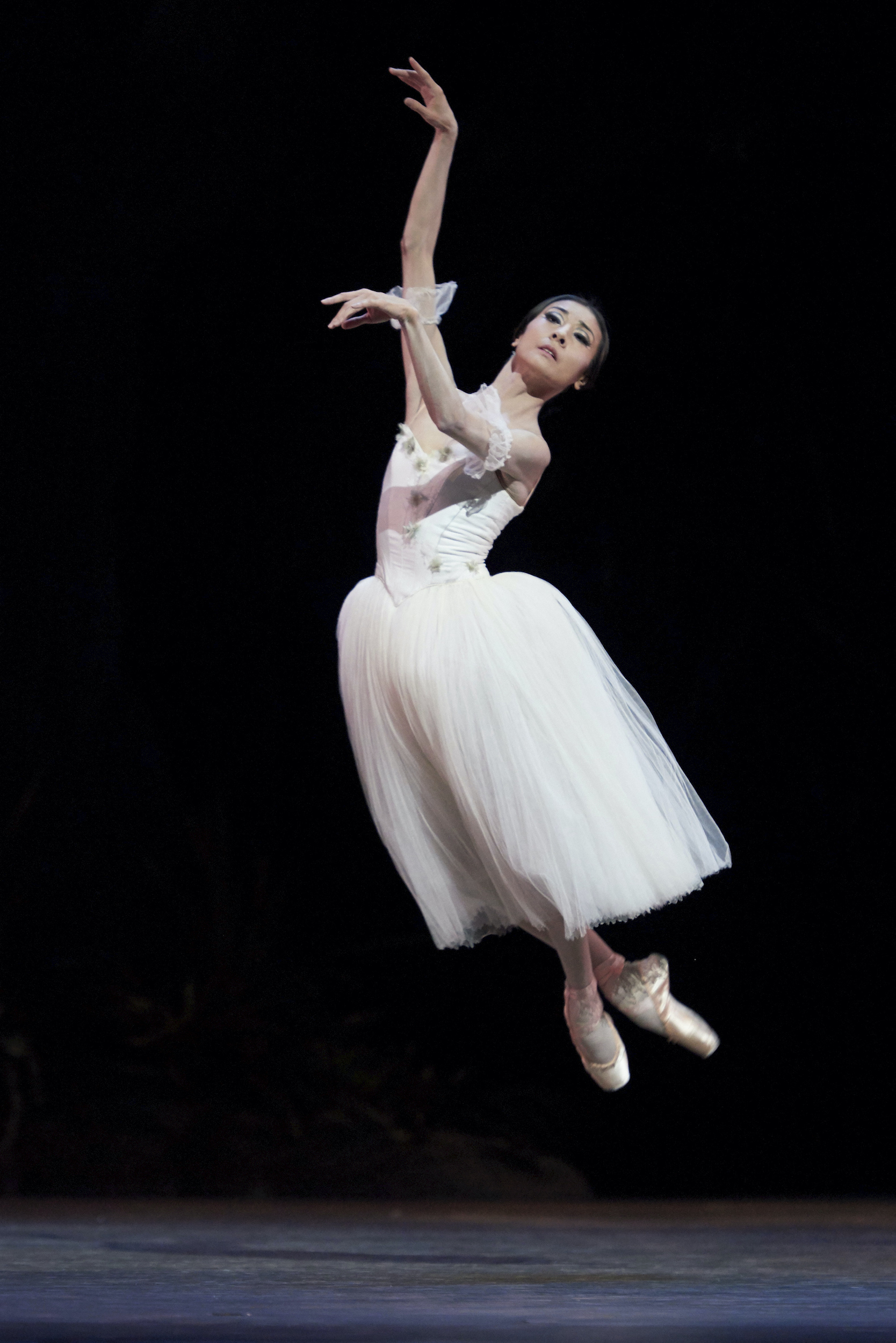 Yuan Yuan Tan jumps up with her crossed feet slightly tucked up underneath her during a performance of Giselle. She lifts her arms to fourth arabesque and looks out over her left hand. Tan wears a long white Romantic tutu, pink tights, and pointe shoes.