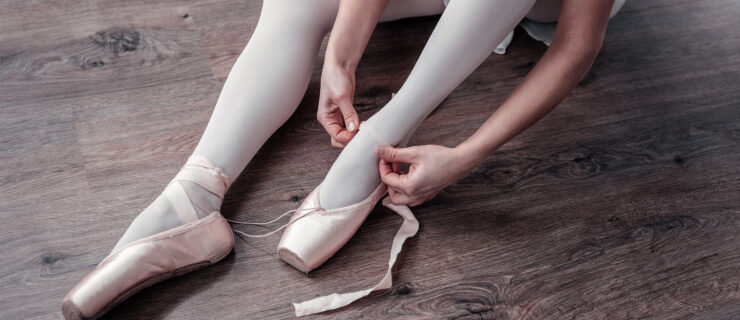 A young woman wearing pink tights and a white skirt is shown from the knees down sitting on a wood floor and putting on a pair of pointe shoes.