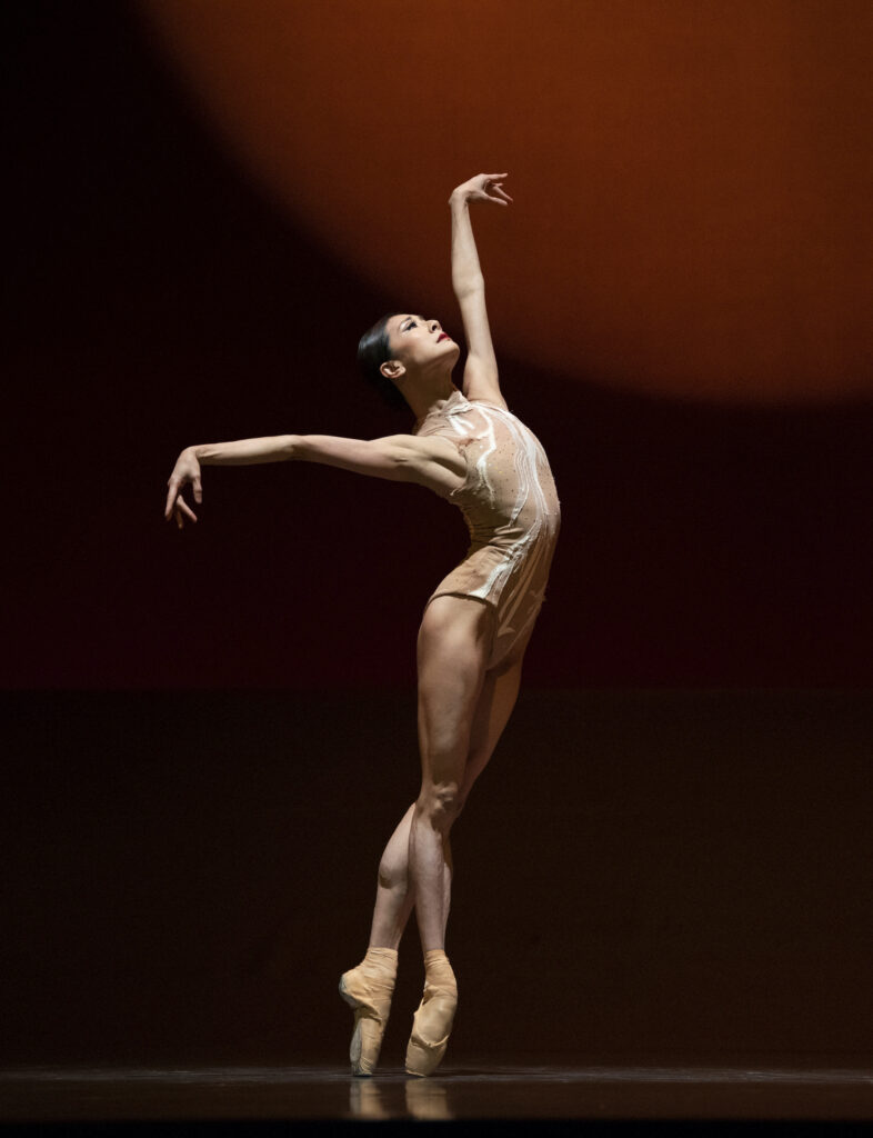 Yuan Yuan Tan stands in a sus-sous on pointe, her body in croisé towards stage left. She leans into her hips slightly and arches back, lifting her left arm up and right arm to the side. She wears a tan colored leotard with a white squiggle design on it, and tan pointe shoes. She performs onstage in front of a dark backdrop with a dark orange spherical projection behind her.