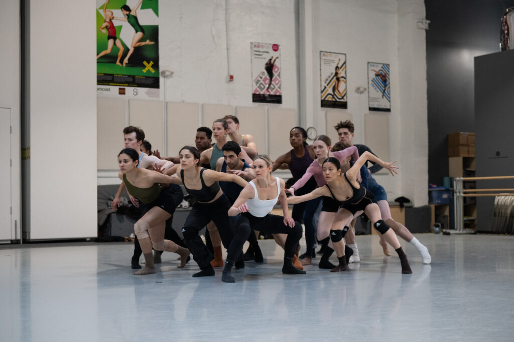 Members of BalletX cluster together in a large group in the center of the studio, reaching out to touch each other while looking away at the downstage left corner.