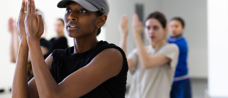Micaela Taylor leading rehearsal at Ballet BC. A close-up shot shows her demonstrating an arm position with backwards-facing prayer hands.