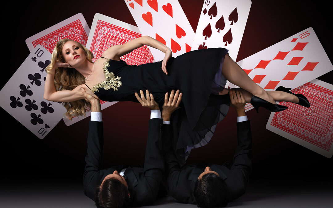 In front of a black backdrop with large suspended playing cards, two men in suits lay on their backs and lift up a woman in a black old-Hollywood gown, who lounges on her side.