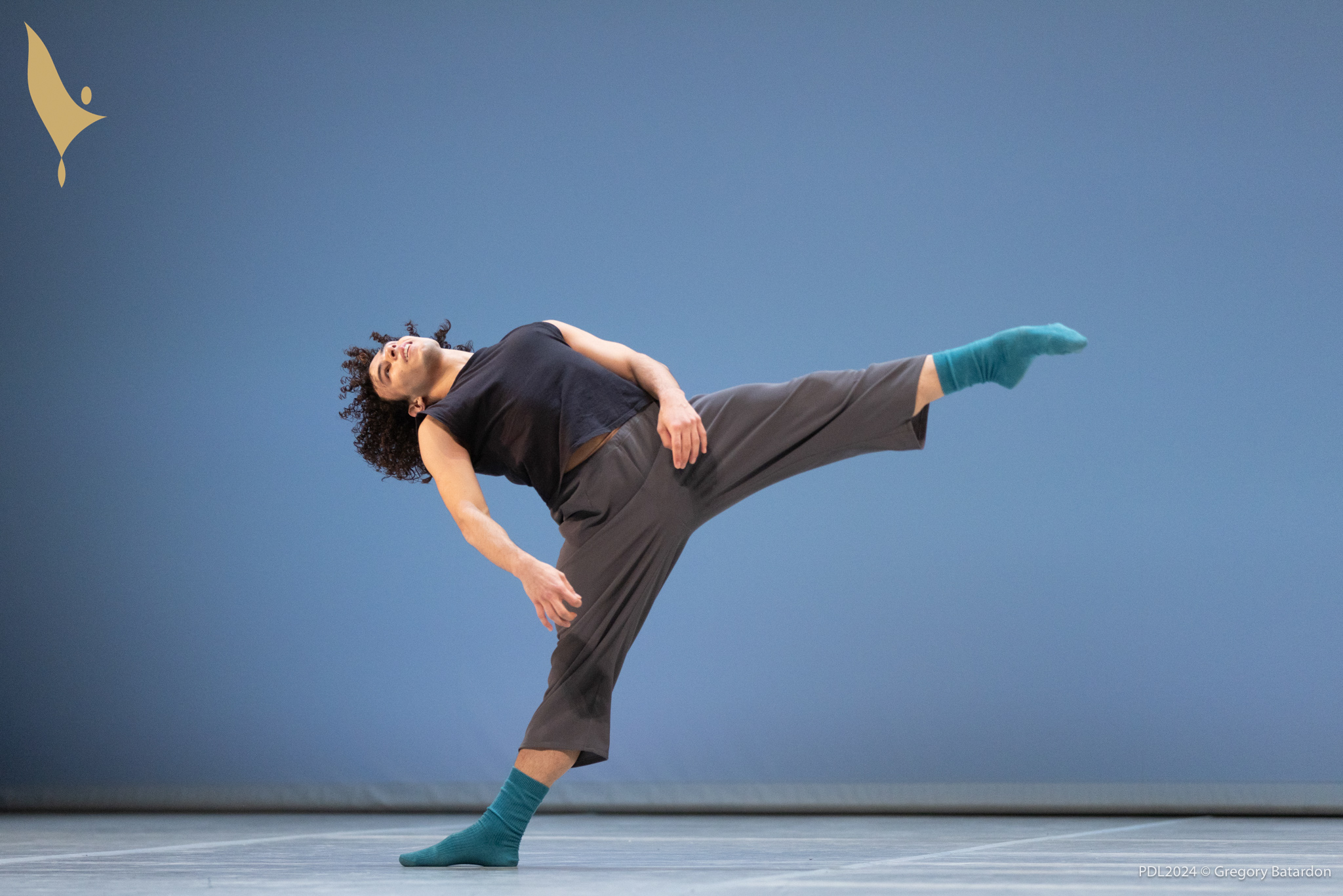 Miguel Artur Alves Oliveira dances onstage, leaning back and doing a layout with his left leg while keeping his arms relazed at his sides. He wears a black sleeveless t-shirt, loose-fitting gray sweatpants and blue socks.