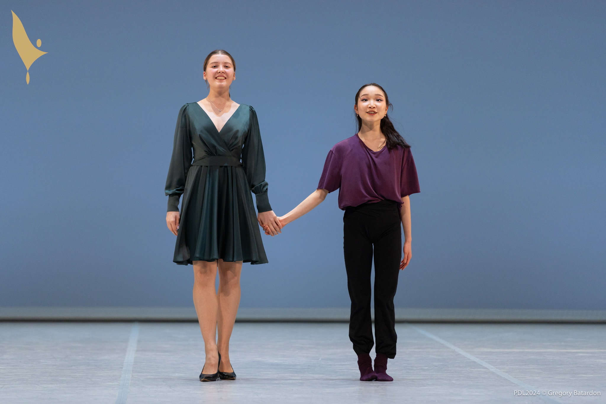 Kseniya Kosava and Sakura Natorigawa hold hands onstage and prepare to take a bow, Kosava wears a short green, long-sleeved dress and heels. Natorigawa wears a purple shirt, black pants and purple socks.