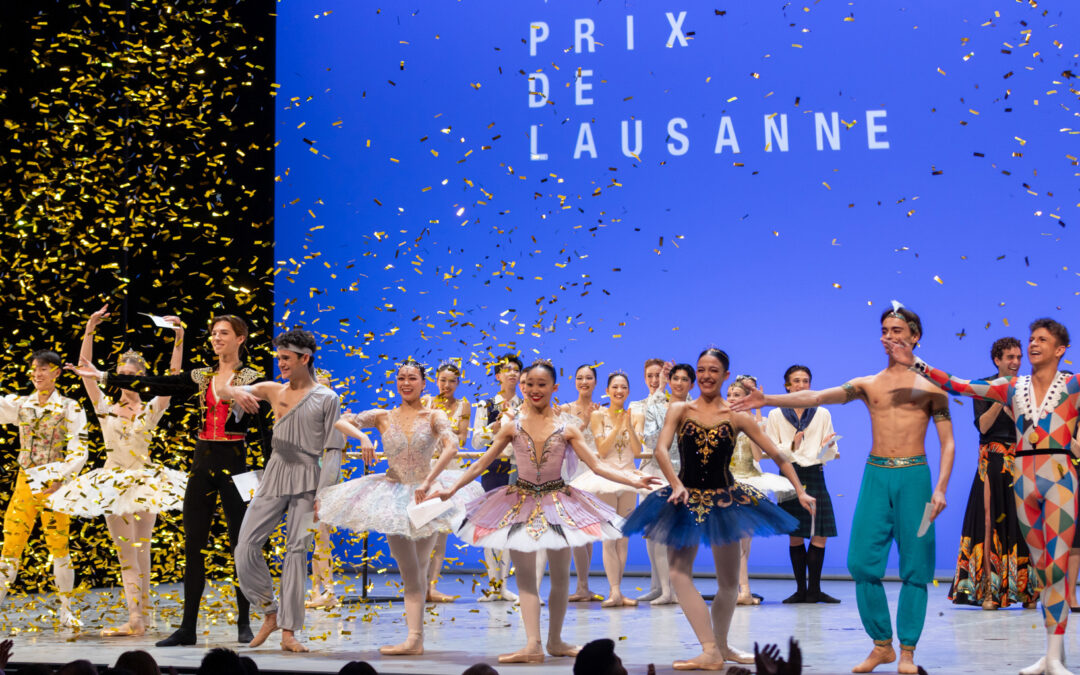 Nine teenage ballet dancers stand in a line at the front of a stage and take a joyful bow. There are four girls and five boys, all dressed in various classical ballet costumes and ballet shoes. Confetti falls from the ceiling. A group of other dancers stand in the background and applaud them.