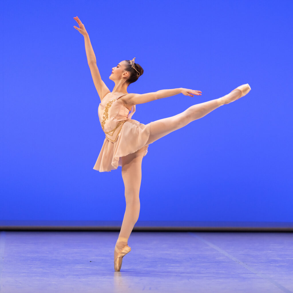 A teenage ballerina performs a first arabesque on pointe during a performance onstage. She is shown in profile facing stage right. She wears a short pink dance dress with gold trim, pink tights and pointe shoes and has a small tiara on her head. She dances in front of a bright blue backdrop.