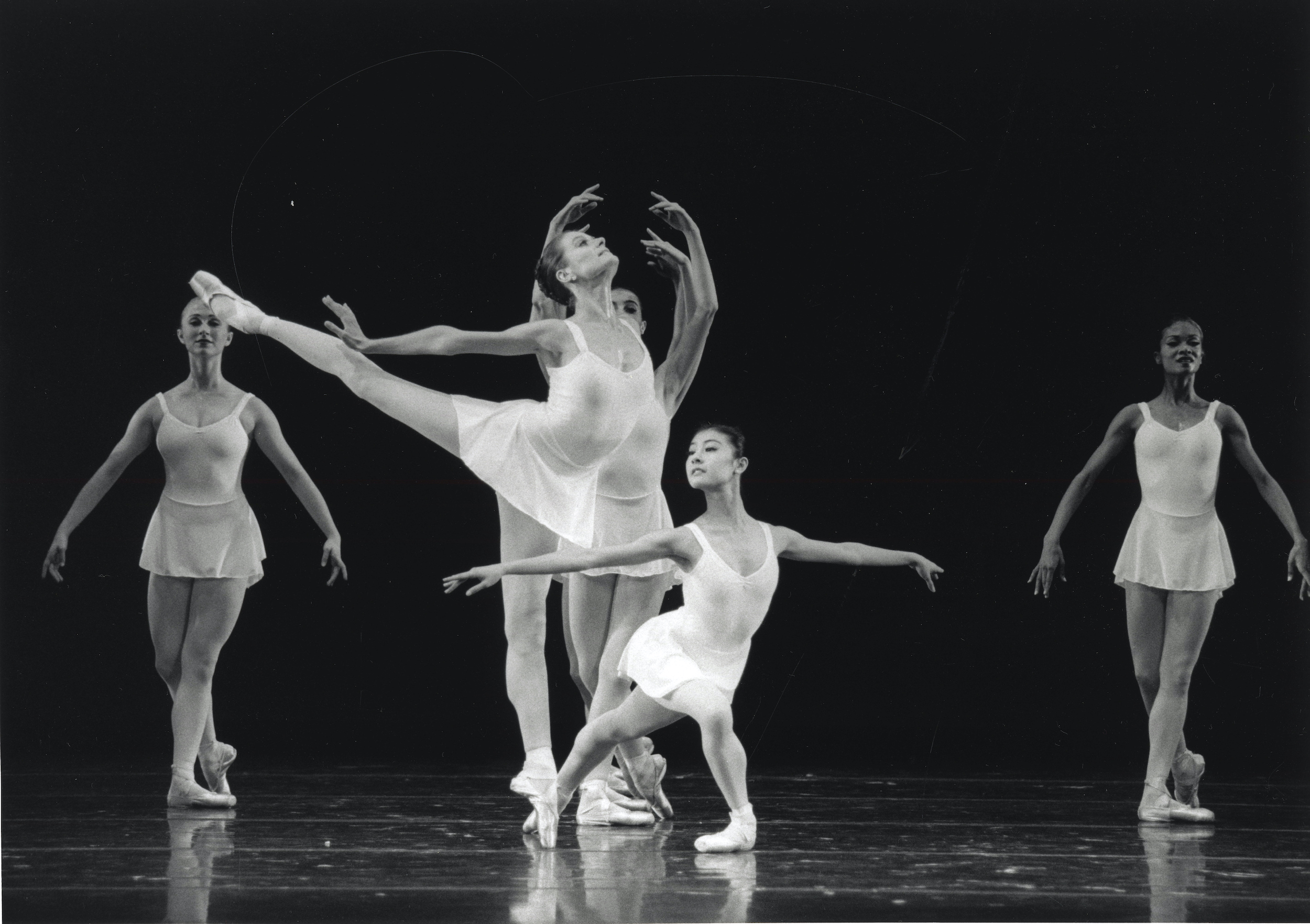 In this black and white photo, YUan YUan Tan lunges low on her right leg, stretching her left leg out diagonally behind her and her arms out to the side as she looks over her right shoulder. Muriel Maffre does an arabesque on pointe next to Tan on her right side, pressing both arms back and looking up. Three women stand in B plus behind them onstage. All of the dancers wear white leotards and short white skirts, tights and pointe shoes.