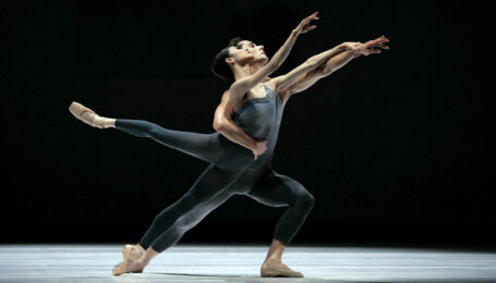 Damian Smith holds Yuan Yuan Tan by the waist with his right arm as she leans forward in a low second arabesque on pointe. They both stretch out their left arms, with Tan holding onto Smith's wrist, and look out wistfully over their hands. They wear dark gray unitards and dance within a spotlight on a darkened stage.