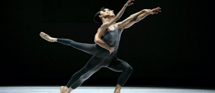 Damian Smith holds Yuan Yuan Tan by the waist with his right arm as she leans forward in a low second arabesque on pointe. They both stretch out their left arms, with Tan holding onto Smith's wrist, and look out wistfully over their hands. They wear dark gray unitards and dance within a spotlight on a darkened stage.