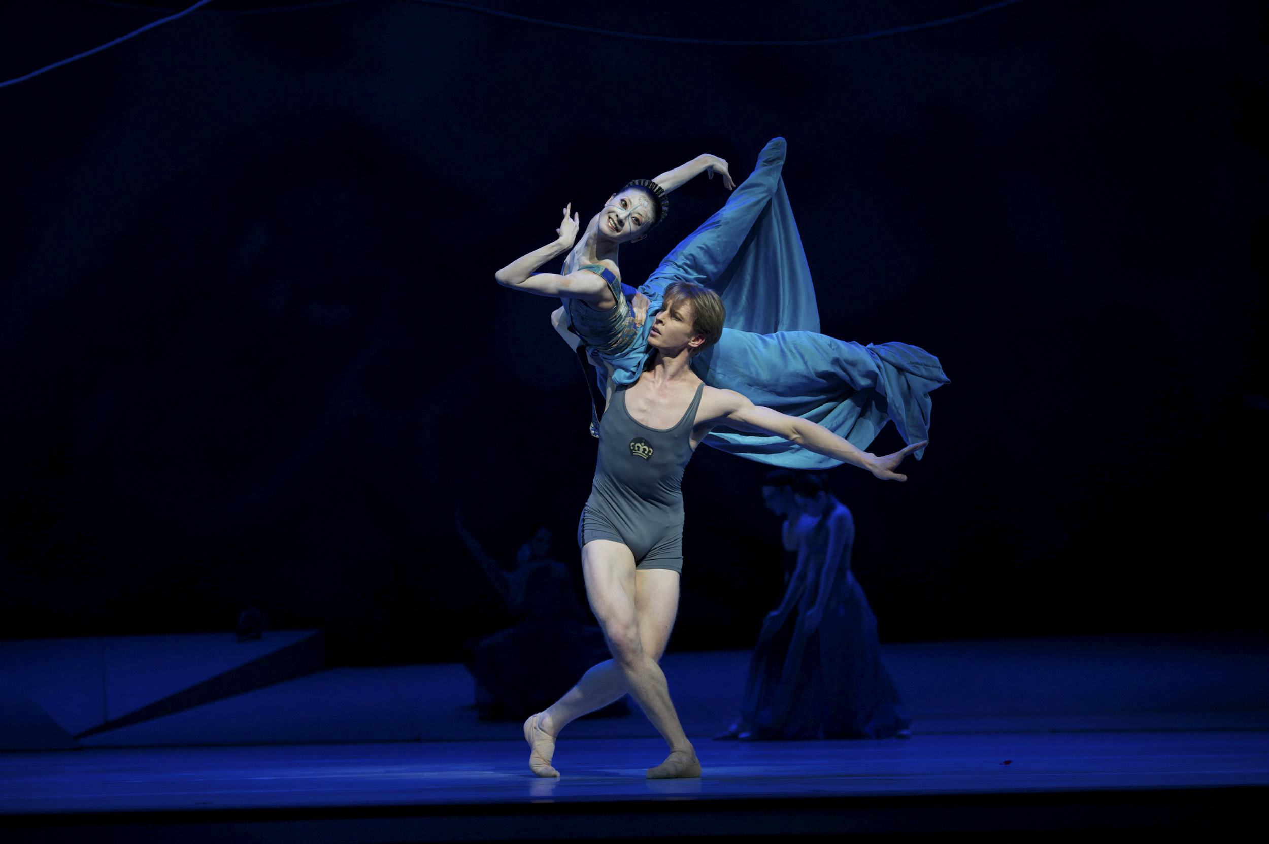 During a performance of The Little Mermaid, Tiit Helimets lifts Yuan Yuan Tan onto his right shoulder. She balances on her hips and extends her legs back, lifting her right leg slightly higher and reaching her right arm back. She bends her left elbow and tucks her left hand under her chin as she looks out into the audience. Helimets wears a turquoise biketard with tank straps. Tan wears a turquoise bodice and long turquoise skirt that wraps around her feet.