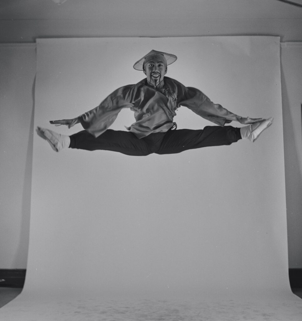 In this black and white photo, George Lee is shown jumping up into the air and splitting his legs open perfectly and reaching to touch his toes. He wears a Chinese-style costume, with black pants, white ballet slippers, a long-sleeved wrap-shirt, and a pointed straw hat.