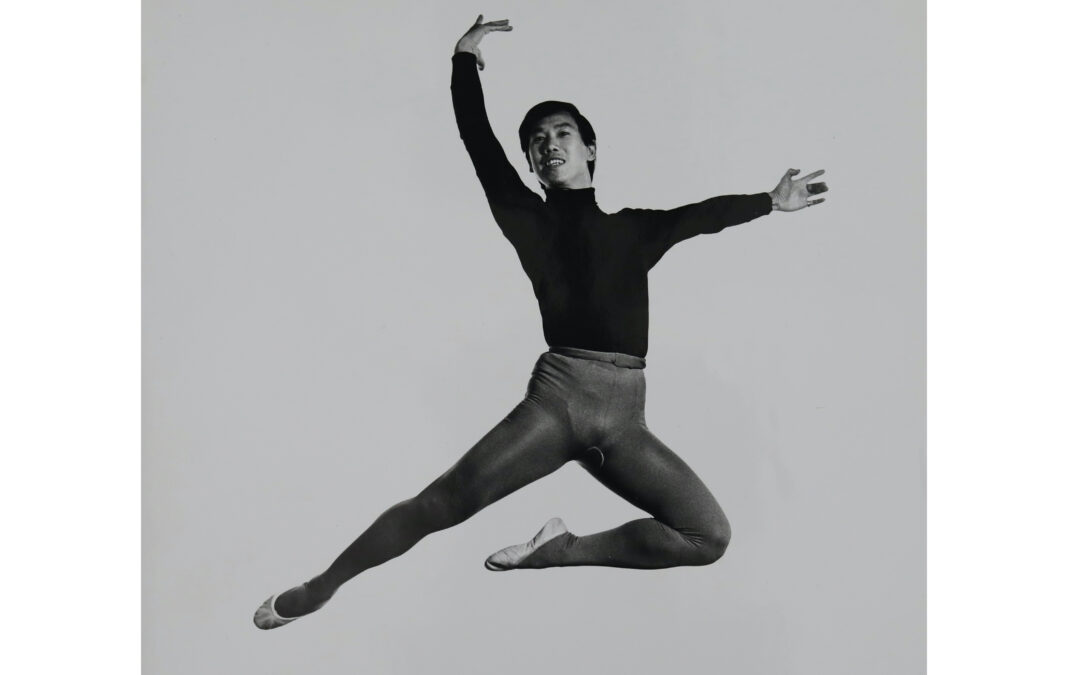 In this black and white photo, George Lee is shown in a black turtle neck, dark tights and white ballet slippers. He does a stag jump, tucking his left leg underneath him, lifting his curved right arm above his head and his left arm out to the side. He looks out toward the camera and smiles.