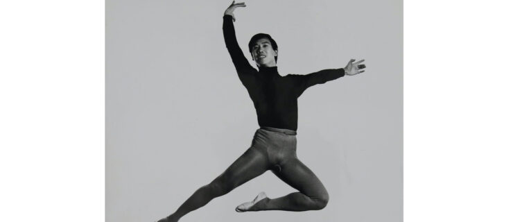 In this black and white photo, George Lee is shown in a black turtle neck, dark tights and white ballet slippers. He does a stag jump, tucking his left leg underneath him, lifting his curved right arm above his head and his left arm out to the side. He looks out toward the camera and smiles.