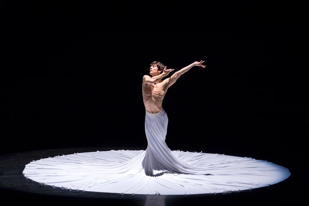 James Yoichi Moore performs in Jessica Lang's <i>TheCalling</i>. He stands onstage in front of a pitch black background in a long, white skirt that creates a huge circle on the floor around him.