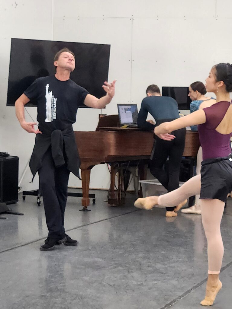In a dance studio, Timour Bourtasenkov gestures with his left hand toward dancer Katherine Cowan.