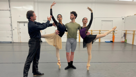 In a dance studio, Timour Bourtasenkov rehearses two female dancers and one male dancer through a pas de trois. Both women stand on pointe in attitude back on either side of the man, who holds them around the waist with each arm.