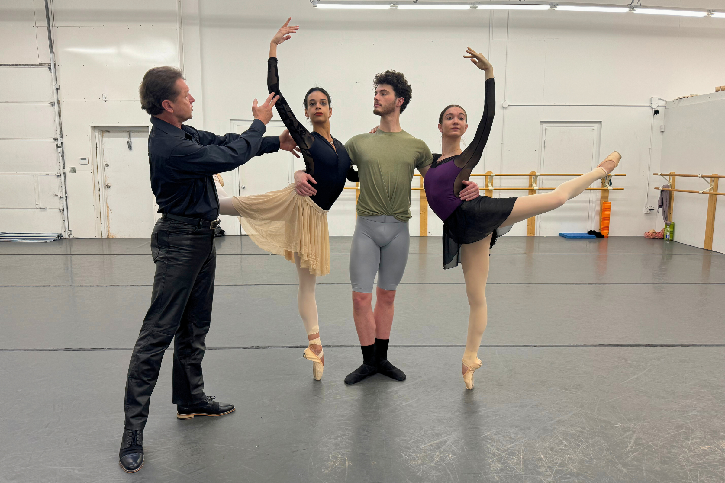 In a dance studio, Timour Bourtasenkov rehearses two female dancers and one male dancer through a pas de trois. Both women stand on pointe in attitude back on either side of the man, who holds them around the waist with each arm.