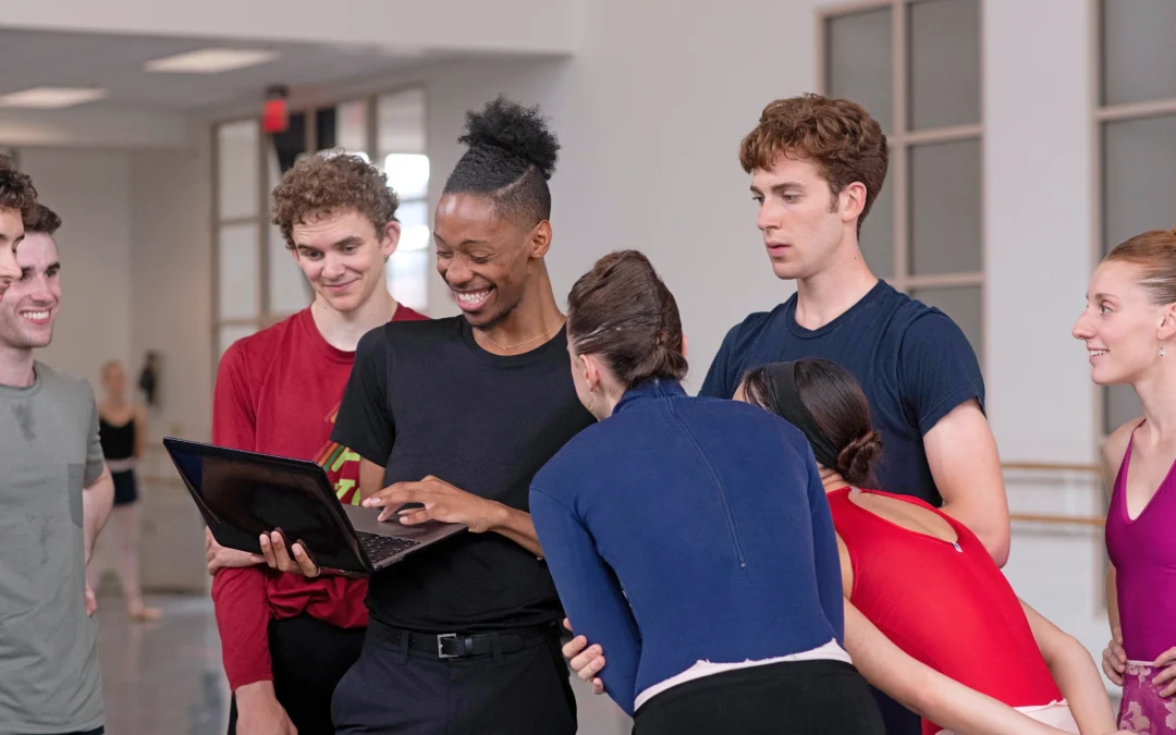 My'Kal Stromile, wearing a black t-shirt and dark pants, holds a laptop in his hands and smiles as a group of male and female dancers in practice clothes surround him and look at what is onscreen. They are in a large dance studio.