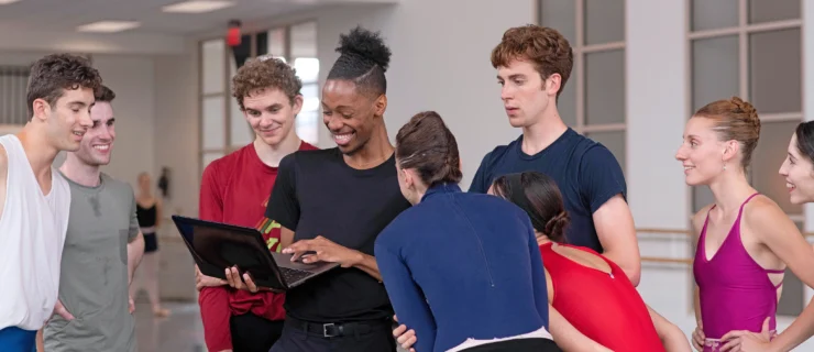 My'Kal Stromile, wearing a black t-shirt and dark pants, holds a laptop in his hands and smiles as a group of male and female dancers in practice clothes surround him and look at what is onscreen. They are in a large dance studio.