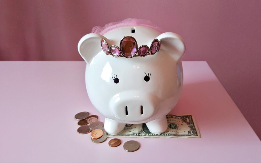 A cute white piggy bank wearing a pink rhinestone tiara and tiny pink tutu rests on top of a pink table. Below its feet is a one dollar bill, and loose change is scattered off to its right side.
