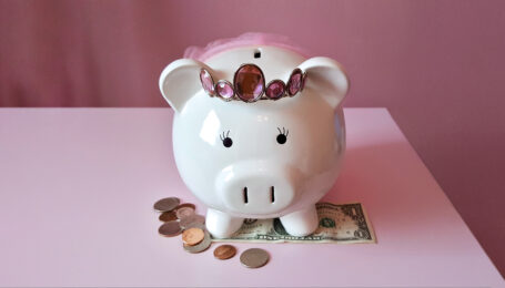 A cute white piggy bank wearing a pink rhinestone tiara and tiny pink tutu rests on top of a pink table. Below its feet is a one dollar bill, and loose change is scattered off to its right side.