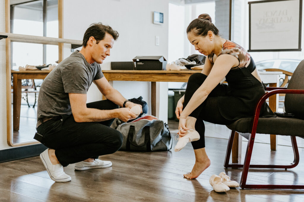 Seth Orza, wearing a gray T-shirt, black jeans and white sneakers, squats down low as an adult female dancer sits in front of him in a chair and tries on a pair of pink canvas ballet slippers. The dancer wears black tights and a black leotard with a multi-colored neck and backline. A wooden table and various bags and boxes are in the background.