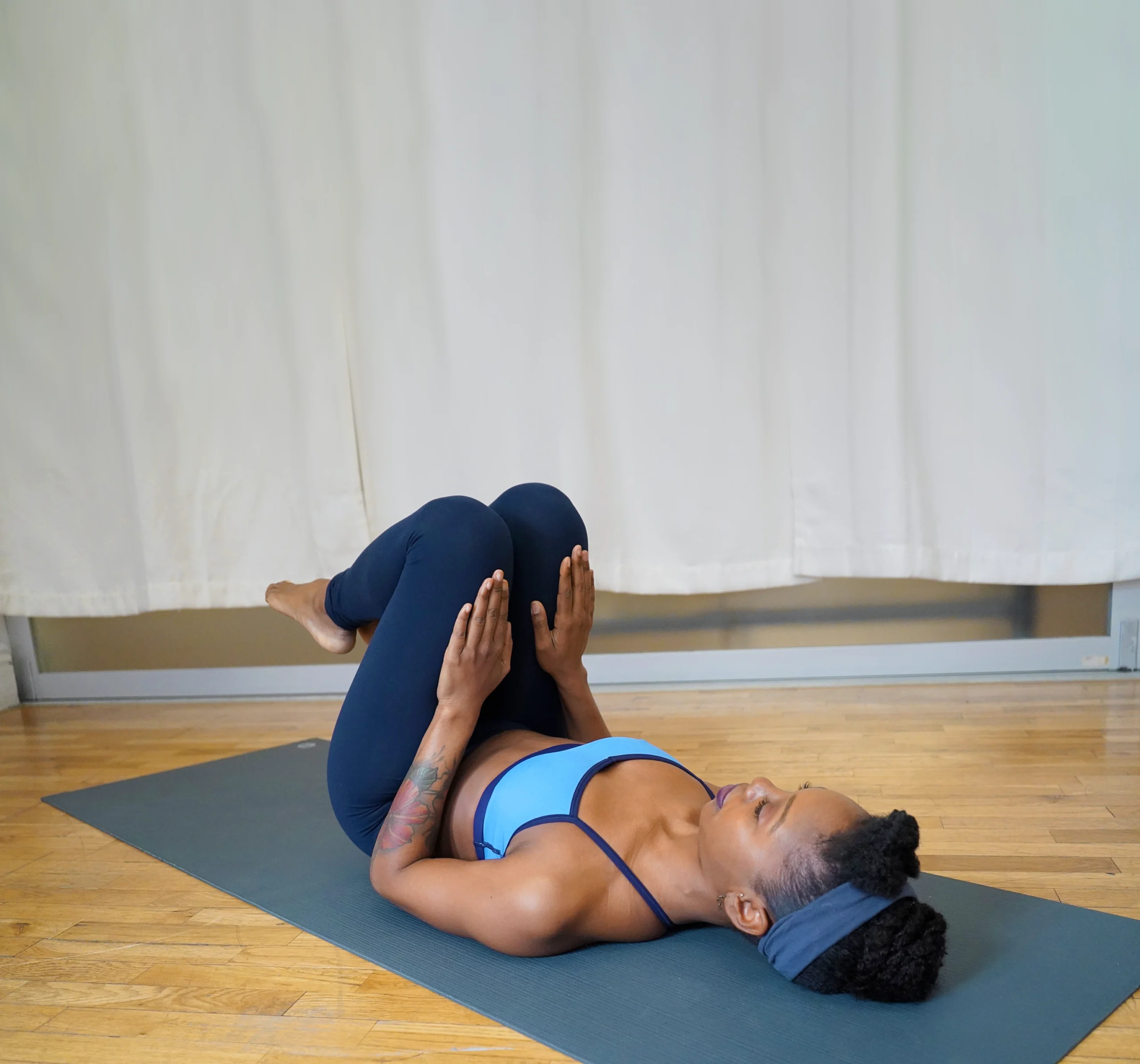A female dancer in dark blue leggings and a light blue sports bra lays on a yoga mat and does an isometric core exercise. She bends both leg in towards her chest and pushes down on her thighs with both palms.