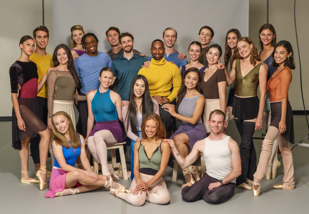 A large group of male and female dancers in various, colorful dance clothing pose for a group photo. They stand or sit close together and smile for the camera.