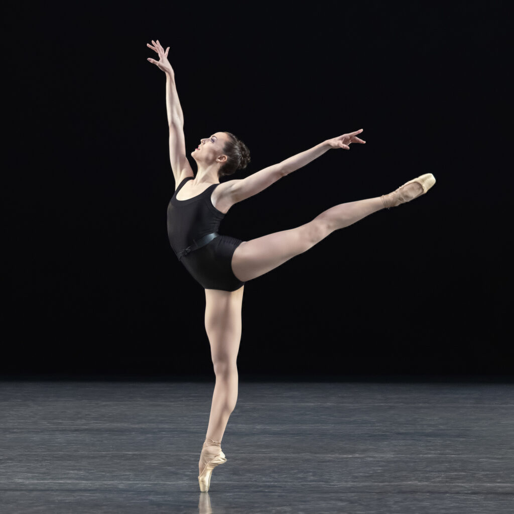 Emily Kikta, costumed in a black leotard, pink tights and pointe shoes, does a huge first arabesque onstage during a performance, facing stage right.