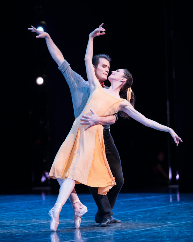 Seth and Sarah Orza dance a romantic pas de deux together onstage. Sarah, wearing a yellow dress, balances in fourth position on pointe with the back leg bent and leans back onto Seth, who ohlds her around the waist. He wearsa a gray shirt and black pants. They look at each other.
