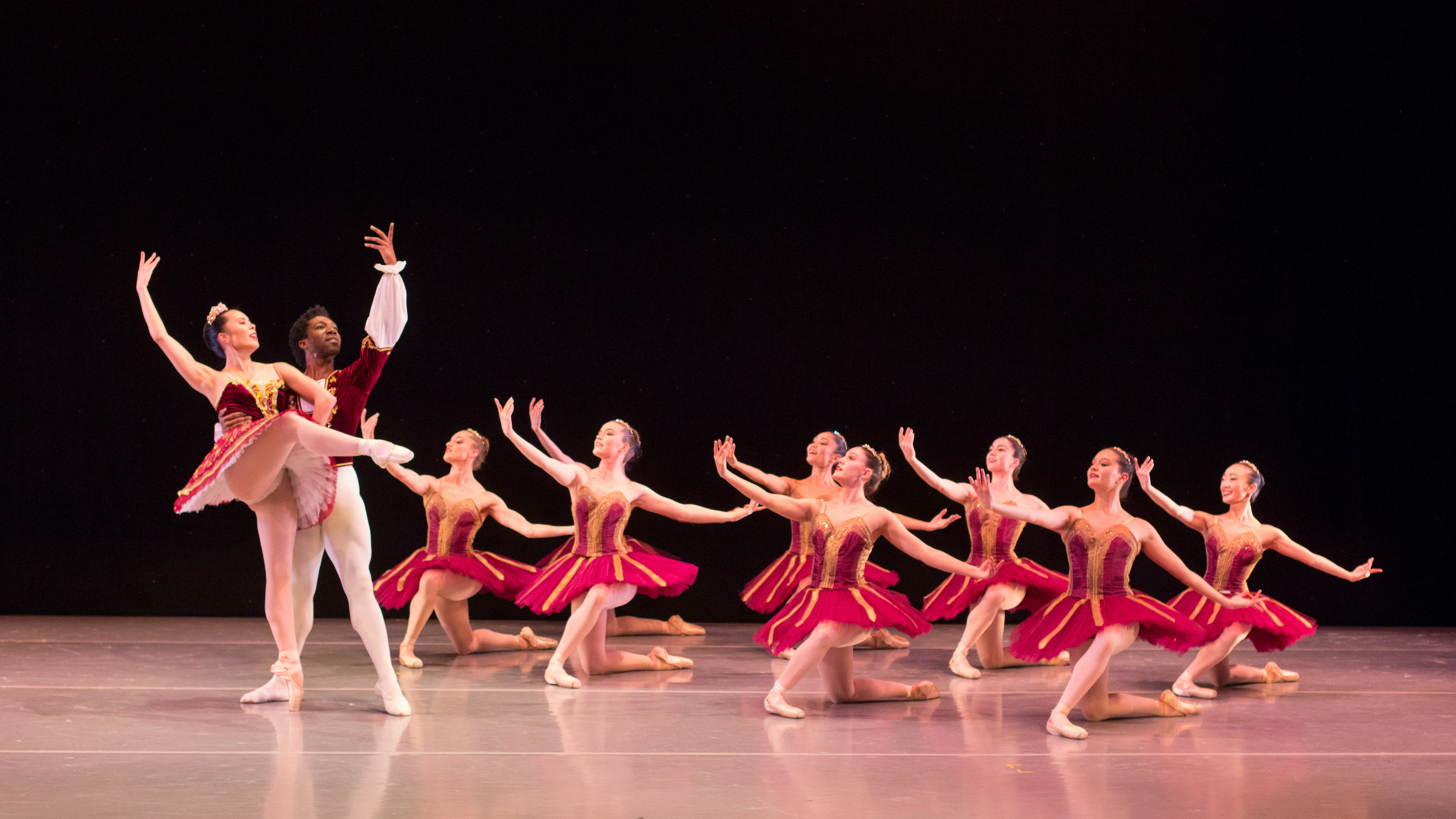 A group of eight ballerinas in red tutus kneel on their right knee in two diagonal lines and gesture toward a lead principal couple to their right. The principal ballerina, in a red tutus and gold tiara, poses in an attitude front on pointe with her right arm raised and her left hand on her hip. Her male partner stands behind her in tendu, holding her waist with his right hand and raising his left arm up.